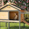 A rabbit is inside a wooden hutch with a ramp leading out. The hutch is set up outdoors on grass, with a fence and red flowers in the background. Produktbild-Vorschau