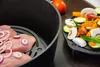 A close-up view of raw meat with onion rings inside a black cooking pot, next to a plate of chopped vegetables and a tomato. Produktbild-Vorschau