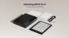 A flat lay of a black e-reader, its packaging box, and some accessories on a light gray background. Produktbild-Vorschau