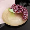 A bunch of red grapes with a leaf rests on a golden, textured plate, which is placed on a light-colored cloth and a black reflective surface. Podgląd zdjęcia produktu