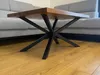 A close-up shot of a coffee table with a wooden top and a black metal spider-leg base, placed on a wooden floor in front of a gray sofa. Anteprima dell’immagine prodotto