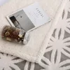 A close-up view of a soft, textured blanket with a decorative pattern, featuring a glass jar filled with potpourri and a magazine. Produktbild-Vorschau