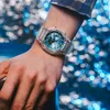 A person wearing a watch on their wrist, with a blurred blue and white background. Anteprima dell’immagine prodotto