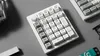 A white mechanical numpad with a rotary knob is placed on a light gray surface, with parts of other keyboards visible in the background. Produktbild-Vorschau