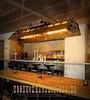A bar interior with a long wooden table in the foreground and a bar with bottles of alcohol in the background. A large, industrial-style pendant light hangs over the table. Produktbild-Vorschau