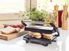 A sandwich maker with two sandwiches inside, placed on a kitchen counter next to some tomatoes and bread. Podgląd zdjęcia produktu