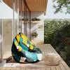 A colorful bean bag chair sits on a wooden deck next to a small woven table, with plants and a stone wall in the background. Náhľad obrázku produktu