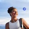 A young woman with headphones on, smiling and looking to the side, with a backpack on her shoulder. The background is a bright blue sky with some clouds and a snowy landscape below. Produktbild-Vorschau
