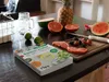 A cookbook is placed on a table with various fruits and a cutting board. Produktbild-Vorschau