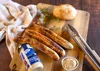 A wooden cutting board with grilled sausages, a bread roll, a jar of mustard, and tongs. Produktbild-Vorschau