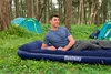 A man is lying on an inflatable mattress in a forest setting with tents in the background. Náhled obrázku produktu