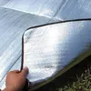 A hand holding up a corner of a silver picnic blanket with a black edge, lying on grass and dirt. Produktbild-Vorschau