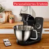 A kitchen mixer with a whisk attachment in a stainless steel bowl, placed on a wooden countertop. A smartphone is visible in the foreground. Produktbild-Vorschau