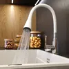 A modern kitchen sink with a white flexible faucet spraying water into the basin. Jars of food items and wooden bowls are visible in the background. Produktbild-Vorschau