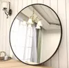 A round mirror with a black frame is displayed on a wooden surface against a white paneled wall. A wall sconce and a chandelier are reflected in the mirror. Anteprima dell’immagine prodotto
