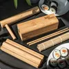 A collection of bamboo sushi-making tools and finished sushi rolls are arranged on a dark surface, with some green plant leaves visible in the background. Náhľad obrázku produktu