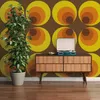 A plant sits next to a mid-century modern dresser with a record player on top, in front of a retro-patterned wallpaper. Produktbild-Vorschau