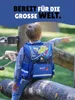 A young boy wearing a backpack is sitting on a wooden beam outdoors, pointing forward. Produktbild-Vorschau