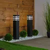 Two black bollard lights are placed in a garden setting, surrounded by white stones, green bushes, and a wooden fence. Anteprima dell’immagine prodotto