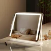 A vanity mirror with integrated LED lighting is placed on a reflective surface, with a bedroom scene reflected in the mirror. Two perfume bottles are visible on the right side. Produktbild-Vorschau