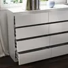 A white dresser with several drawers, some open, against a window with greenery visible outside. Voir les images du produit