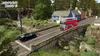 A video game screenshot showing a rural Scottish landscape with a bridge, a river, a waterfall, houses, a black car, and a red double-decker bus. Produktbild-Vorschau