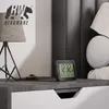 A digital alarm clock is placed on a nightstand next to a lamp. Podgląd zdjęcia produktu