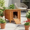 A wooden cat house with a cat sleeping inside, placed on a patio with potted plants and outdoor furniture. Produktbild-Vorschau