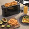 A black toaster with four slices of toast is shown on a kitchen counter, alongside toast with avocado and tomatoes, a glass of orange juice, and cut avocados. Produktbild-Vorschau
