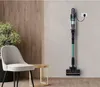 A cordless vacuum cleaner is mounted on a wall and charging, next to a chair and a small table with a plant. Produktbild-Vorschau