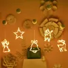 Christmas-themed window decorations with LED lights, including a bell, star, angel, and reindeer, hanging against an orange wall with decorative paper flowers. Podgląd zdjęcia produktu