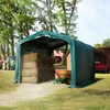 A green tarpaulin shelter with hay bales and a small orange and green trailer inside, with a tractor and a red truck in the background. Voir les images du produit