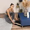 A woman in workout clothes is placing a vibration plate under a blue coffee table. Náhľad obrázku produktu