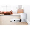 A white robot vacuum cleaner is docked at its charging station next to a white wall, with a blurred living room in the background. Produktbild-Vorschau