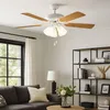 A white ceiling fan with wooden blades and lights is shown above a living room setting with a couch and bookshelves. Anteprima dell’immagine prodotto