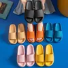 Person, die schwarze Schlapfen hält, mit Paaren von orangen, beigen, blauen, pinken und gelben Schlapfen auf blauer Fläche angeordnet Anteprima dell’immagine prodotto