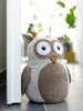 A stuffed owl doorstop is placed next to a white door frame on a floor. Plants are visible in the blurred background. Anteprima dell’immagine prodotto