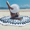 Rundes Strandtuch mit blau-weißem geometrischen Muster und Fransensaum, auf Sand ausgelegt, mit einer Person mit weißem Hut Anteprima dell’immagine prodotto