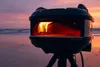 A portable pizza oven is set up on a tripod on a beach at sunset. Produktbild-Vorschau