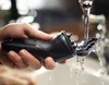 A hand is holding a black electric shaver under running water from a faucet, cleaning it. Podgląd zdjęcia produktu