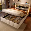 A bedroom scene with a bed that has storage compartments open, showing folded items inside. The bed has a built-in headboard with shelves and lighting. Náhľad obrázku produktu