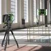 A laser level device is set up on a tripod in a large, well-lit indoor space with tall windows and columns. A person in a safety vest is visible in the background, and a magnified view of the device's display is shown in a callout box. Anteprima dell’immagine prodotto