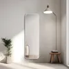 A full-length mirror is placed against a wall next to a potted plant and a floor lamp. A small wooden stool with folded towels sits beside the mirror. Voir les images du produit