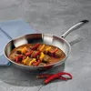 A stainless steel frying pan with food inside, placed on a textured gray surface with a blue cloth and red chilies nearby. Podgląd zdjęcia produktu