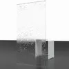 A clear glass shower screen with water droplets on it, standing upright on a dark surface, with a white stool-like object behind it. Produktbild-Vorschau