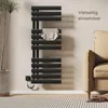 A black electric towel warmer is mounted on a light-colored wall, with a towel draped over one of its bars. A potted plant is to the left, and a portion of a beige sofa is visible to the right. The floor has a striped rug. Anteprima dell’immagine prodotto