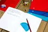 An open notebook with handwritten text, a pencil, and a protractor on a wooden surface, with other notebooks and gummy bears in the background. Produktbild-Vorschau