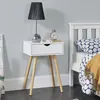 A white bedside table with a drawer and wooden legs, next to a bed. Náhled obrázku produktu