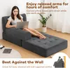 A woman is sitting on a gray sofa bed, using a laptop. The sofa is in a living room setting with a plant and a rug. Produktbild-Vorschau