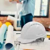 A white construction helmet is placed on top of blueprints, with rolled-up blueprints and other construction-related items in the foreground. A person is out of focus in the background. Produktbild-Vorschau
